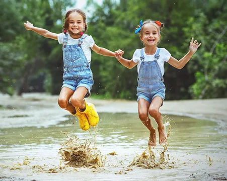 Two twin sisters jump in a muddy puddle.