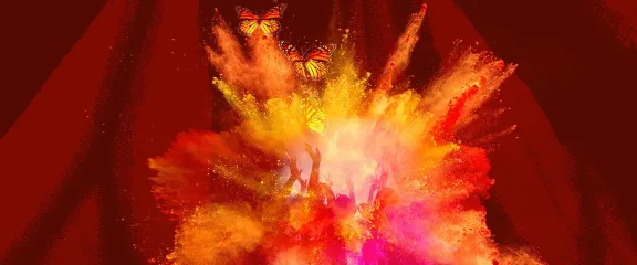 A colourful explosion of dust in yellows, oranges and reds with the raised hands on dancers behind it and butterflies in front