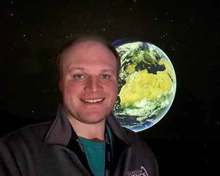 Planetarium presenter, Hayden, smiles while standing in front of the Earth displayed on the fulldome